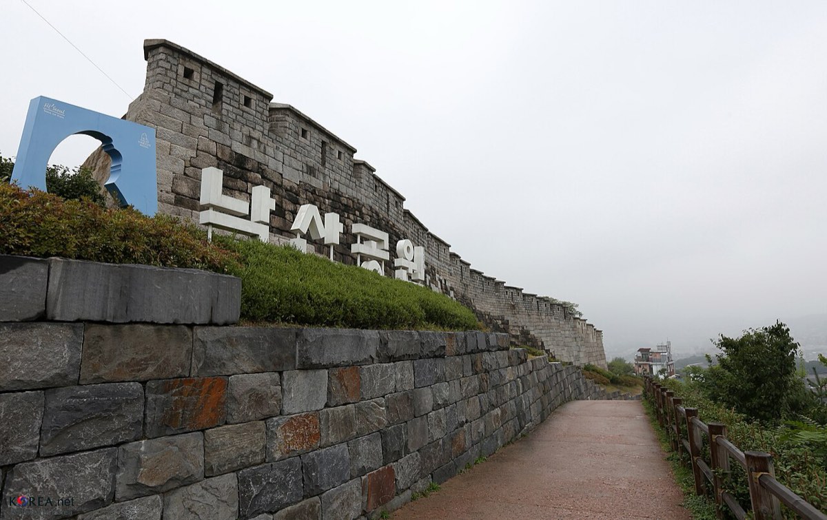 The Seoul City Wall climbing Naksan beside the park sign, with a walking path along its base.