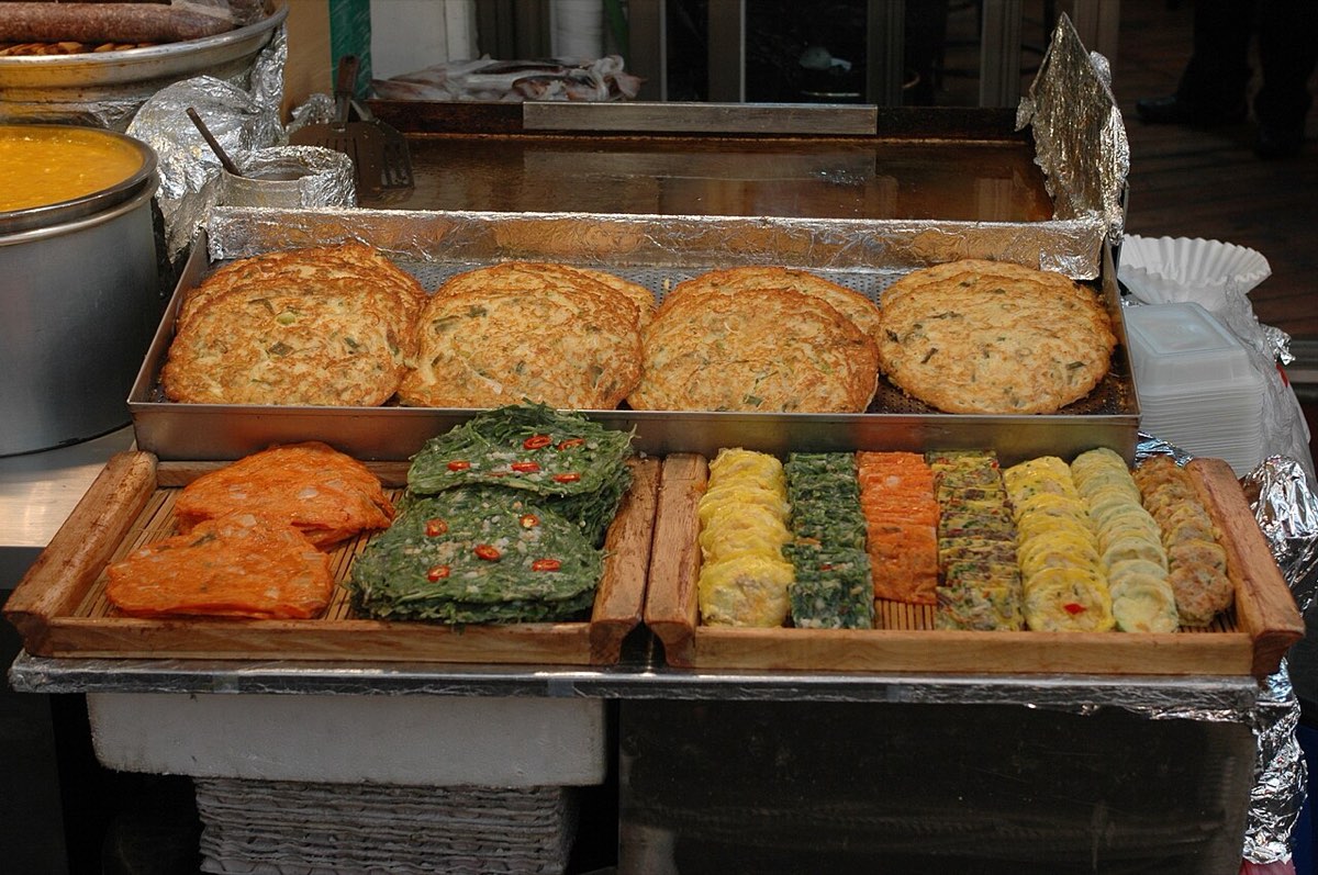 Assorted jeon, pan-fried pancakes, vegetables and fish, laid out on trays along a Gwangjang Market alley.