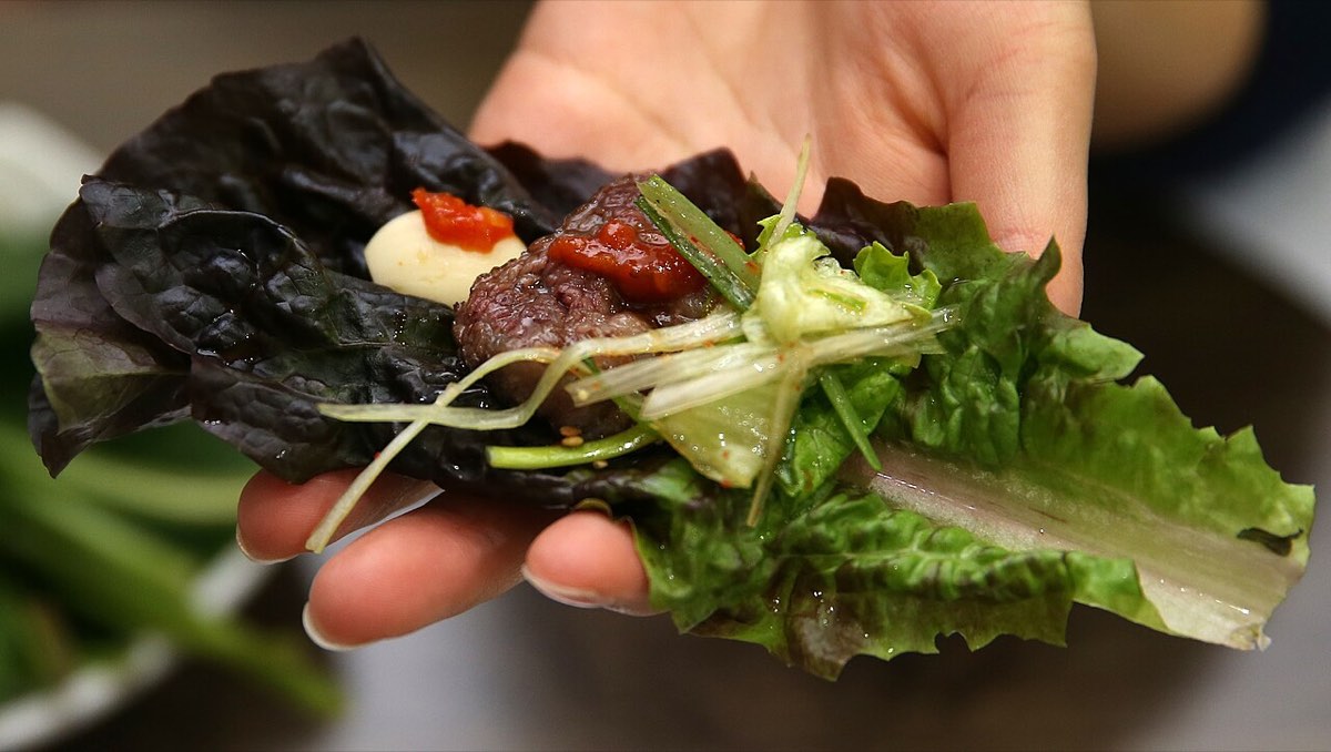 A hand holds a lettuce ssam wrapped around a slice of grilled beef with garnishes.