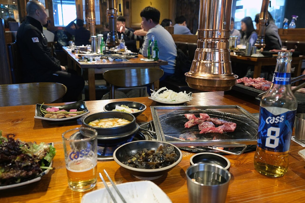 A Korean barbecue table in Seoul with meat on the grill, side dishes, a glass and a bottle of beer.