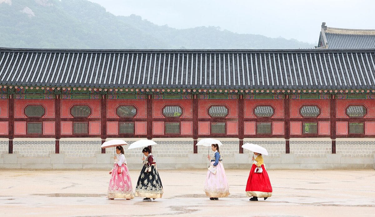 Visitors in hanbok carry white umbrellas past a palace wall at Gyeongbokgung during Seoul's rainy season.